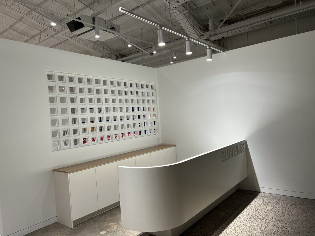 A modern white reception desk with "GLOBEWEST" signage stands in front of a white wall featuring a grid of small framed images, creating an inviting space where reliable support awaits. Ceiling track lights provide bright, even illumination.