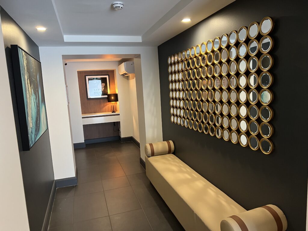 A hallway with a beige bench, a wall art installation of round mirrors, dark floor tiles, and a framed artwork near a well-lit vanity area in the background.